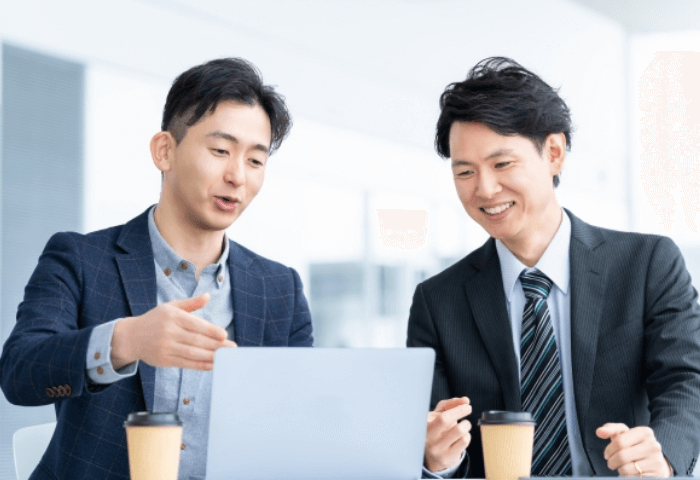 男性2名が笑顔で話している画像
左の男性が右の男性にPCを見ながらアドバイスをしている場面