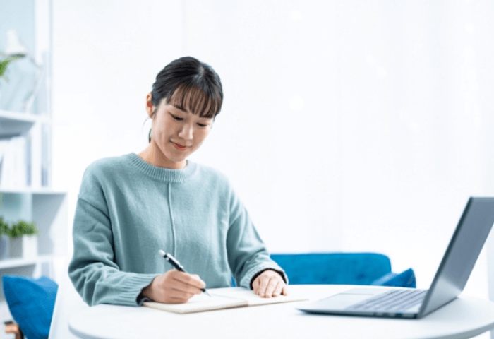 ノートパソコンで動画講義を受けながらメモを取って勉強している女性の画像